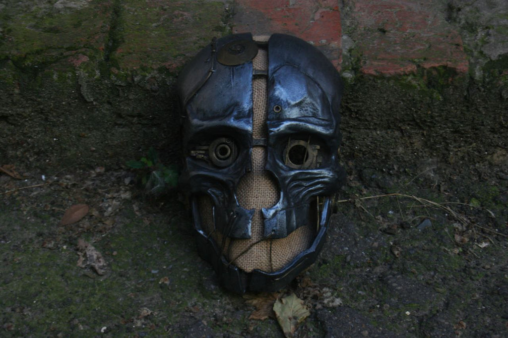 A wearable cosplay mask inspired by Dishonored Corvo, featuring a black and silver design with straps, placed on a surface with a brick wall in the background.